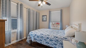 Une chambre à coucher comprenant un lit, un ventilateur de plafond, une fenêtre avec des stores et une table de chevet avec une lampe.