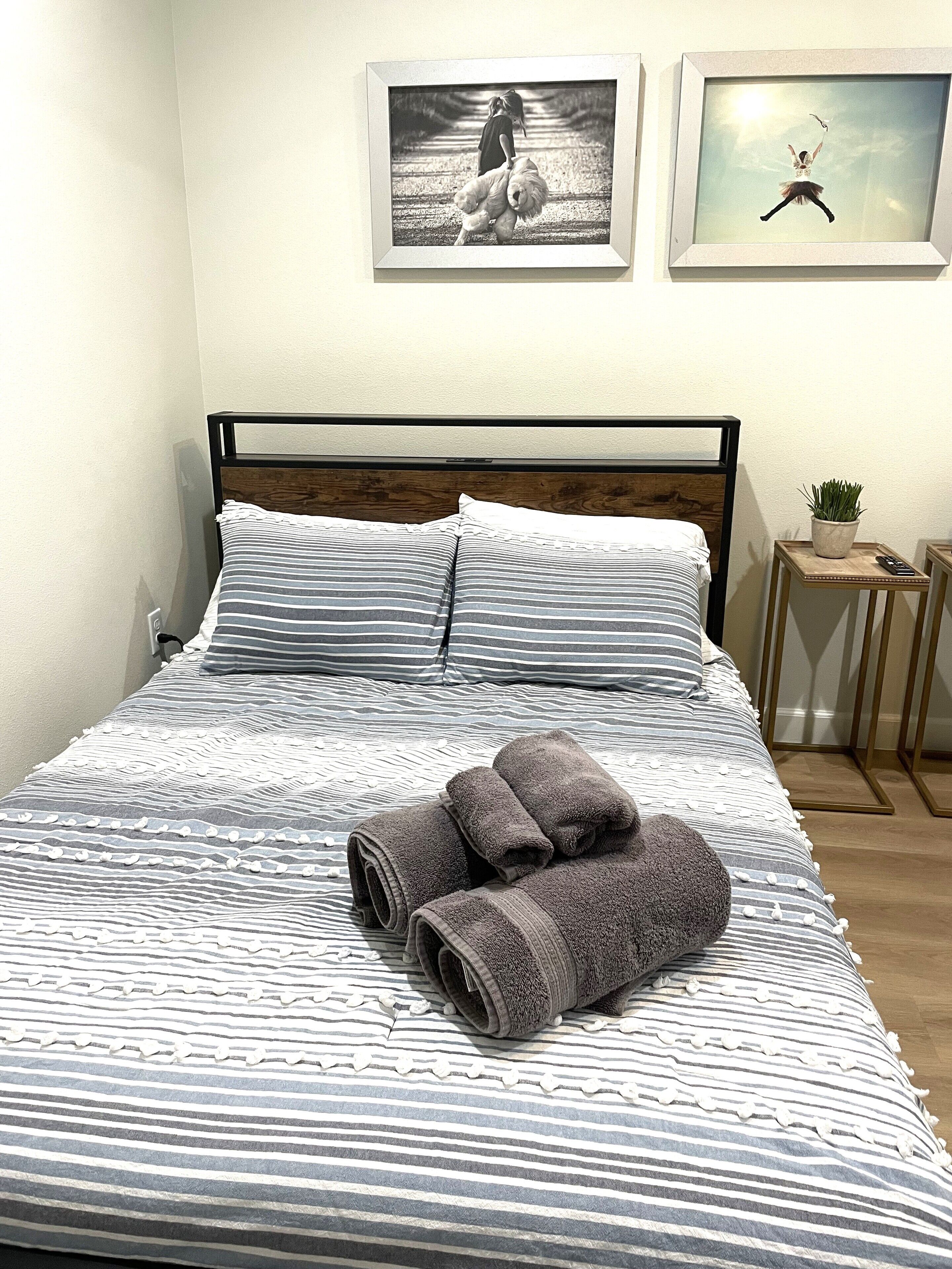 Een net opgemaakt bed met gestreept beddengoed, een paar opgerolde handdoeken en twee ingelijste foto's aan de muur.
