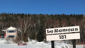 A sign reading "Le Remous '18" in front of a house with a snowy landscape and a forest in the background.
