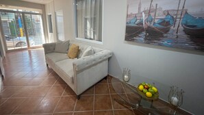 A living room with a sofa, a glass coffee table with fruit, a painting of boats, and a view through a window. - Shorebreak Hotel (Aguadilla)