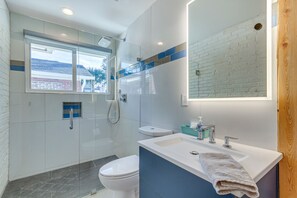 Ein modernes Badezimmer mit einer Duschkabine aus Glas, einer weißen Toilette, einem Waschbecken mit Handtuch und einem Spiegel, der eine Ziegelwand reflektiert.