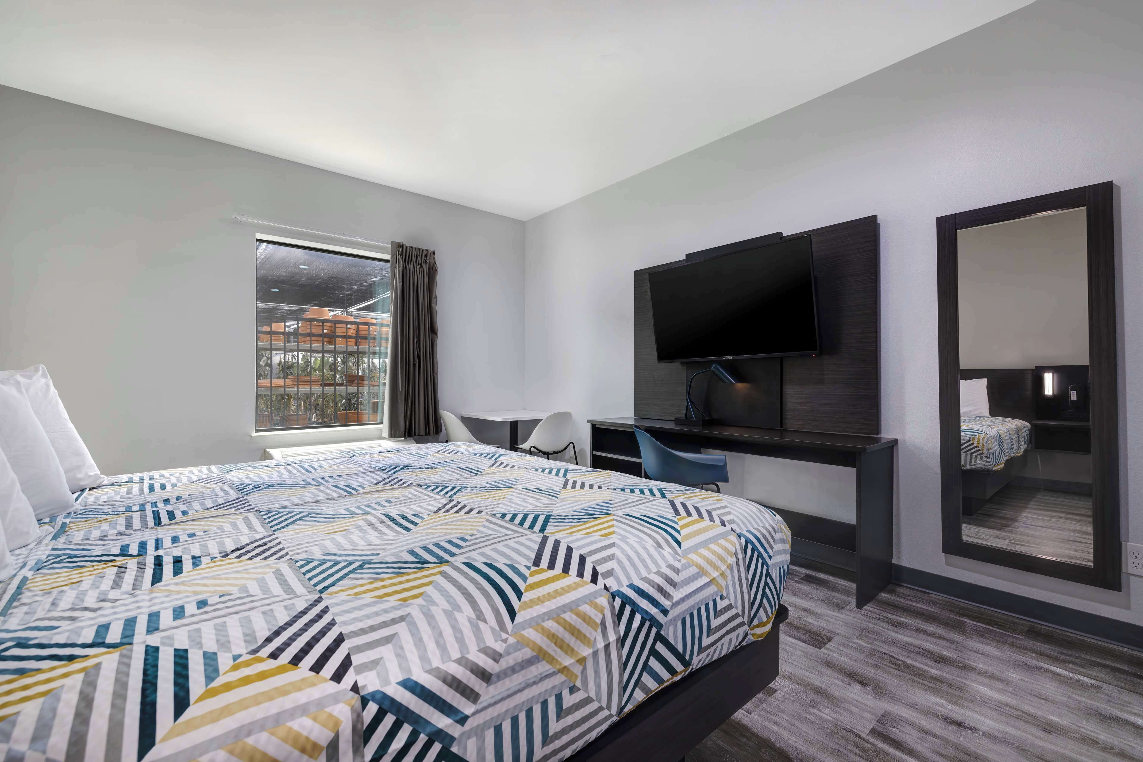 A modern hotel room with a large bed, a desk, a flat-screen TV, a mirror, and a window with a view of buildings.