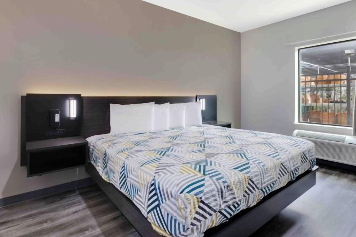 A modern hotel room with a large bed, bedside tables, a nightstand, a window with a view, and a wall-mounted light fixture.