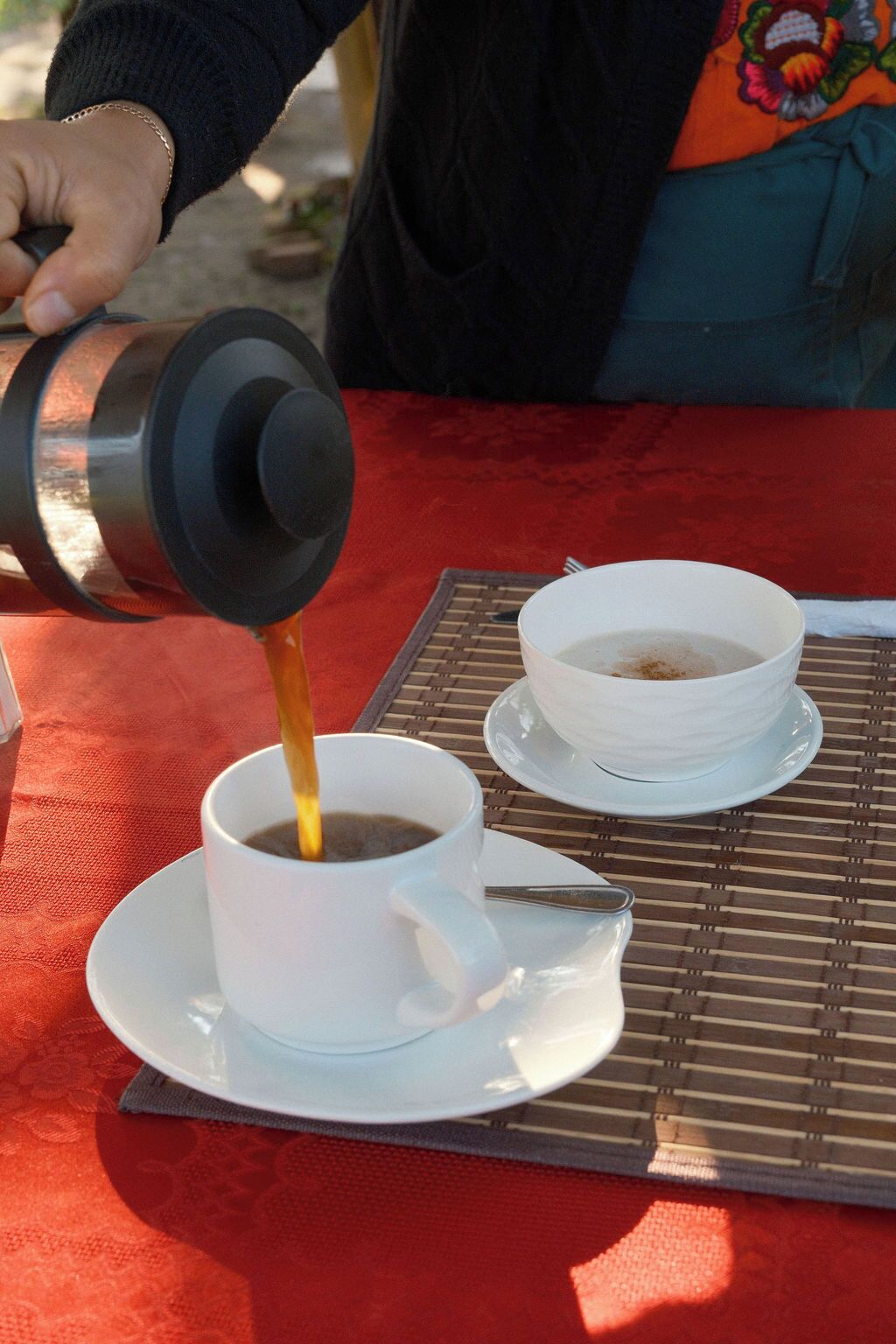A person pouring coffee into a cup on a red tablecloth.
