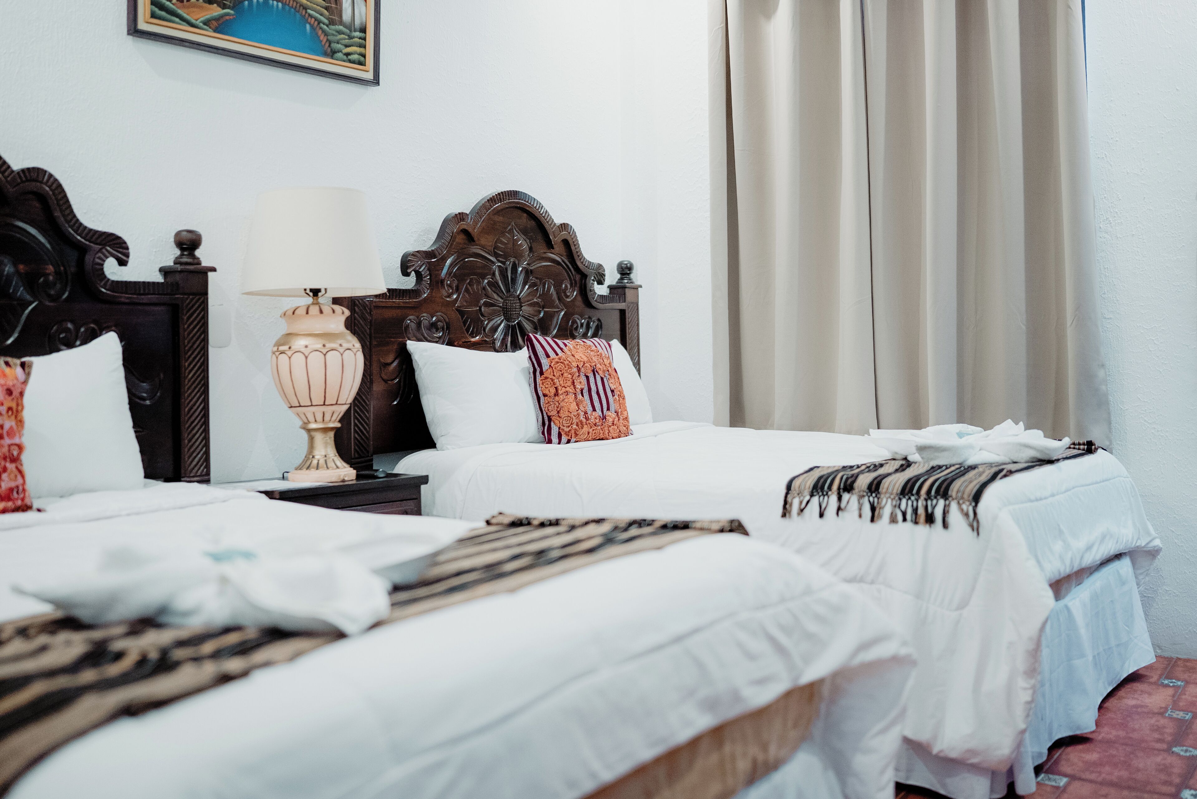 A hotel room with two single beds, a wooden headboard, a bedside table with a lamp, and a painting on the wall.