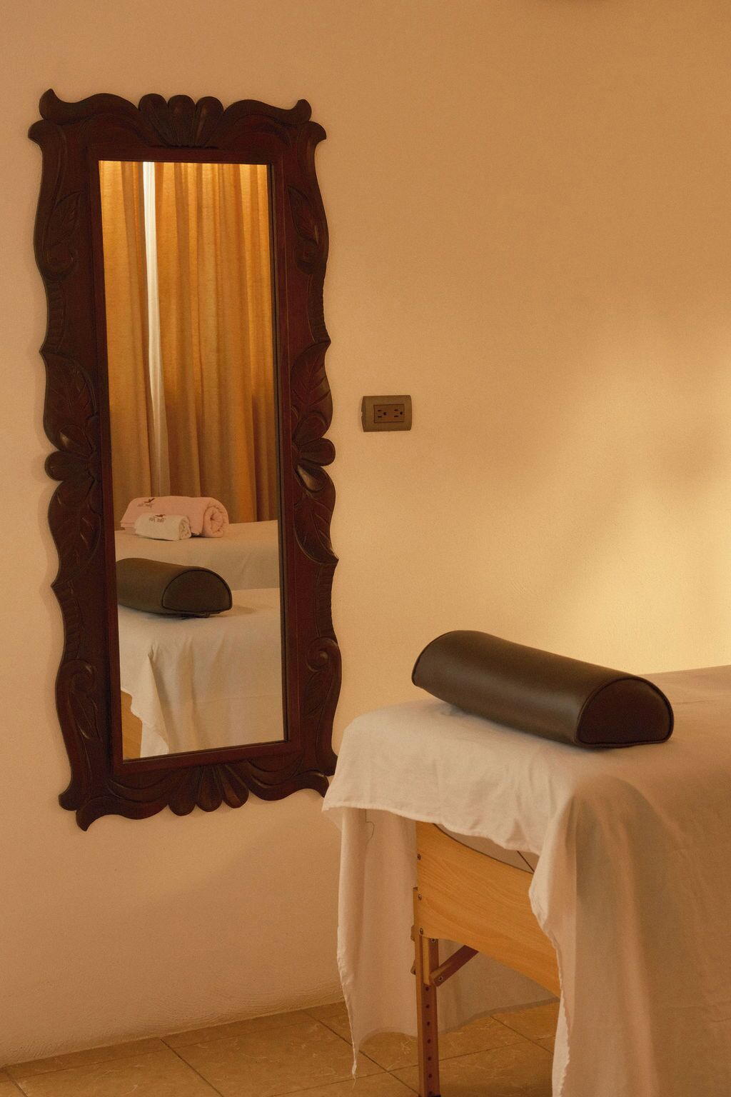 A room with a large mirror, a bed with a white sheet, a pillow, and a bedside table with a black object.