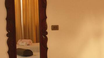 A room with a large mirror, a bed with a white sheet, a pillow, and a bedside table with a black object.