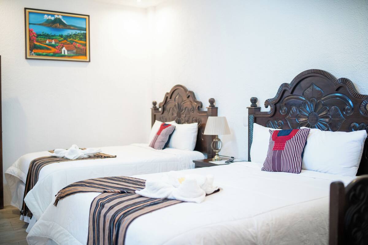 A hotel room with two beds, each with a wooden headboard, white bedding, and striped blankets. There is a painting on the wall above the left bed.