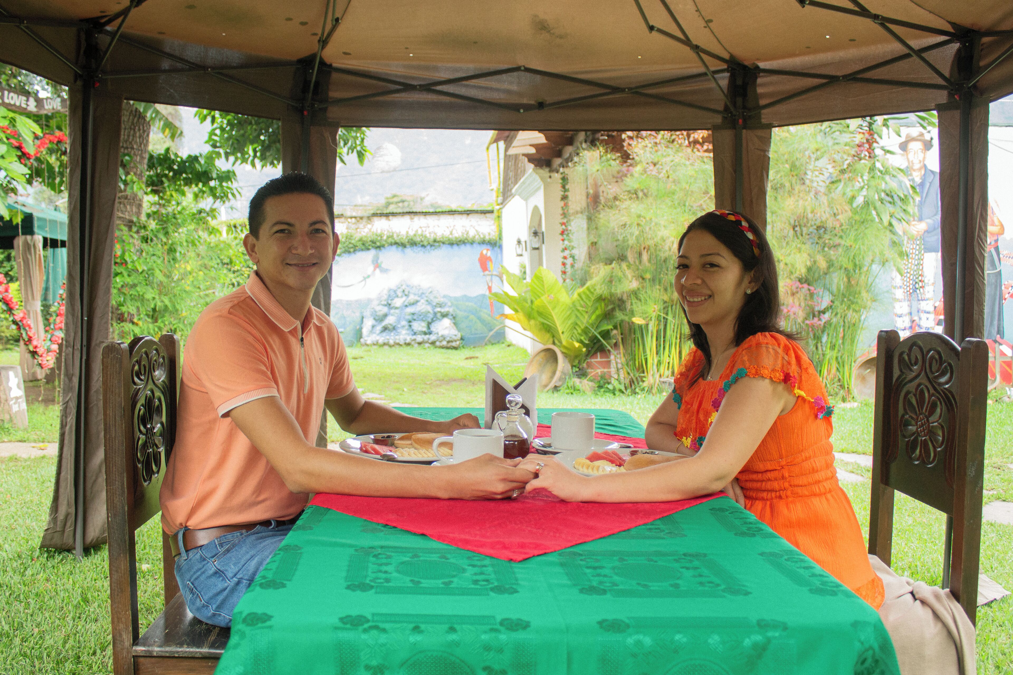 Um casal jantando em uma mesa ao ar livre com toalha de mesa verde, cercado por cadeiras e uma área coberta com vista para um jardim e um edifício ao fundo.