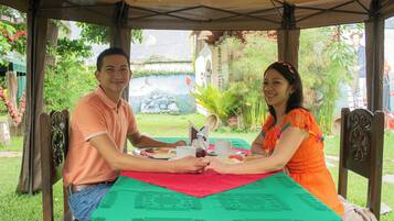 Um casal jantando em uma mesa ao ar livre com toalha de mesa verde, cercado por cadeiras e uma área coberta com vista para um jardim e um edifício ao fundo.