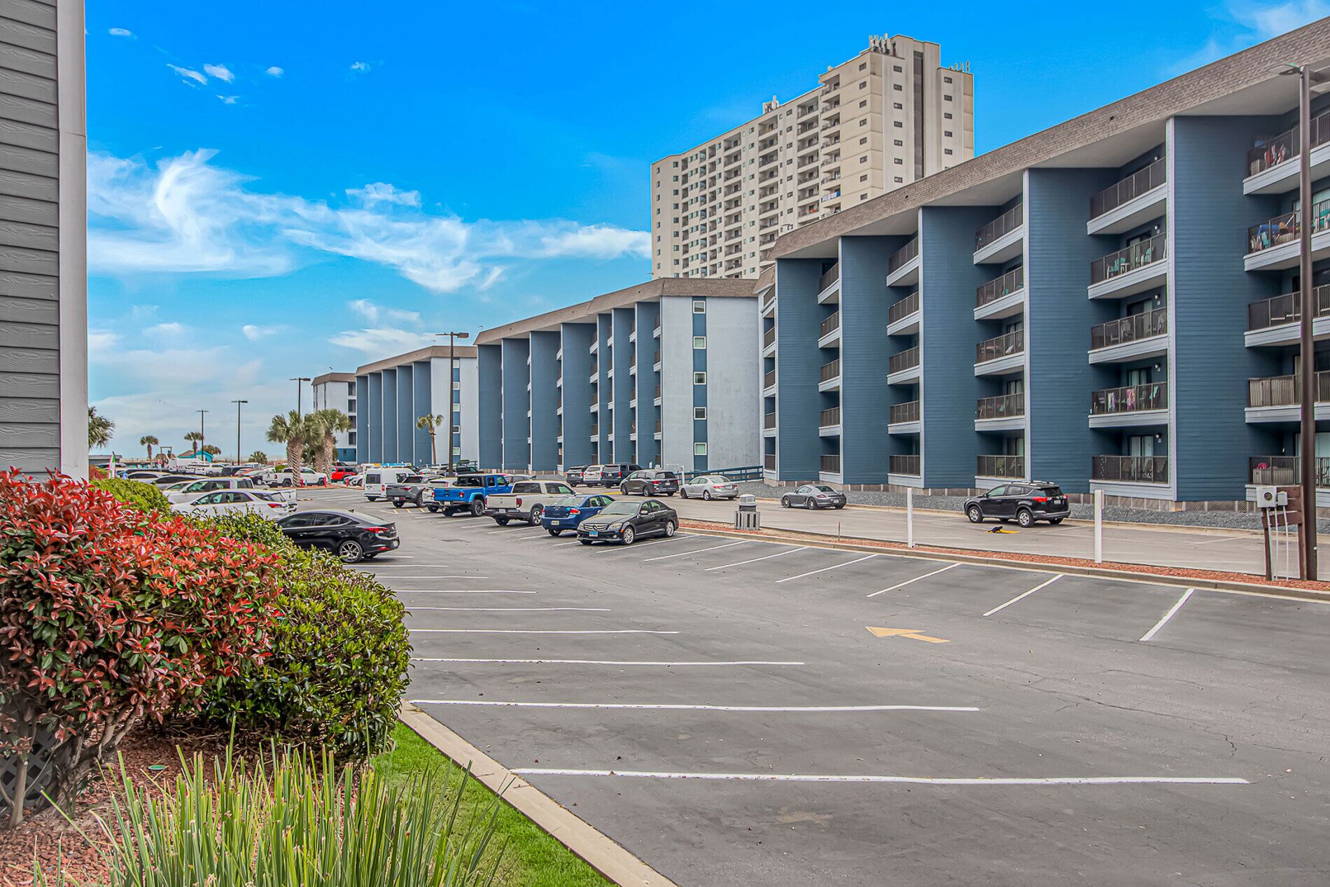 A parking lot with multiple cars, a building with balconies, and a clear blue sky.
