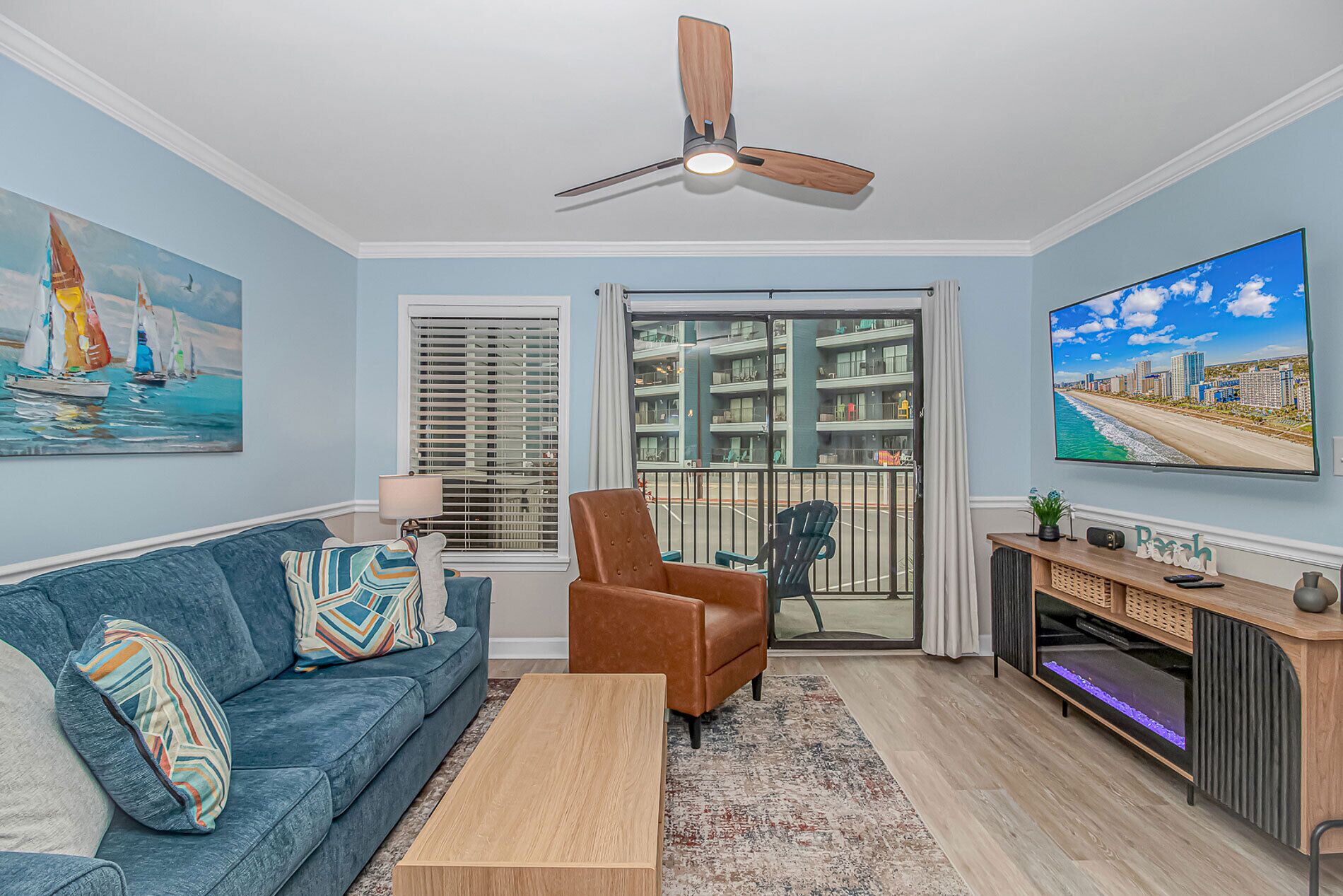 A living room with a blue sofa, a brown armchair, a wooden coffee table, a ceiling fan, a large TV, and a view of a cityscape through a window.