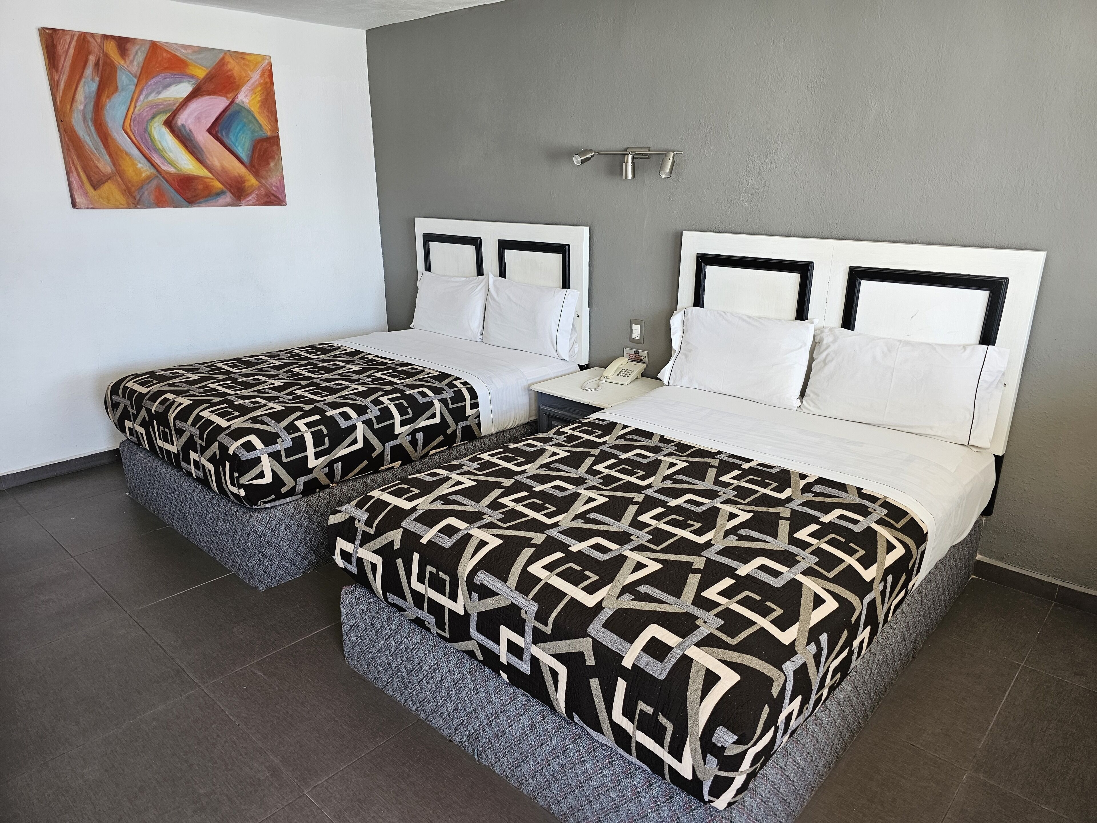A hotel room with two beds, a gray wall, a colorful abstract painting, and a phone on the nightstand.