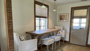 Comedor con mesa de madera y cuatro sillas, ventana con persianas, puerta con panel de vidrio y una lámpara colgante.