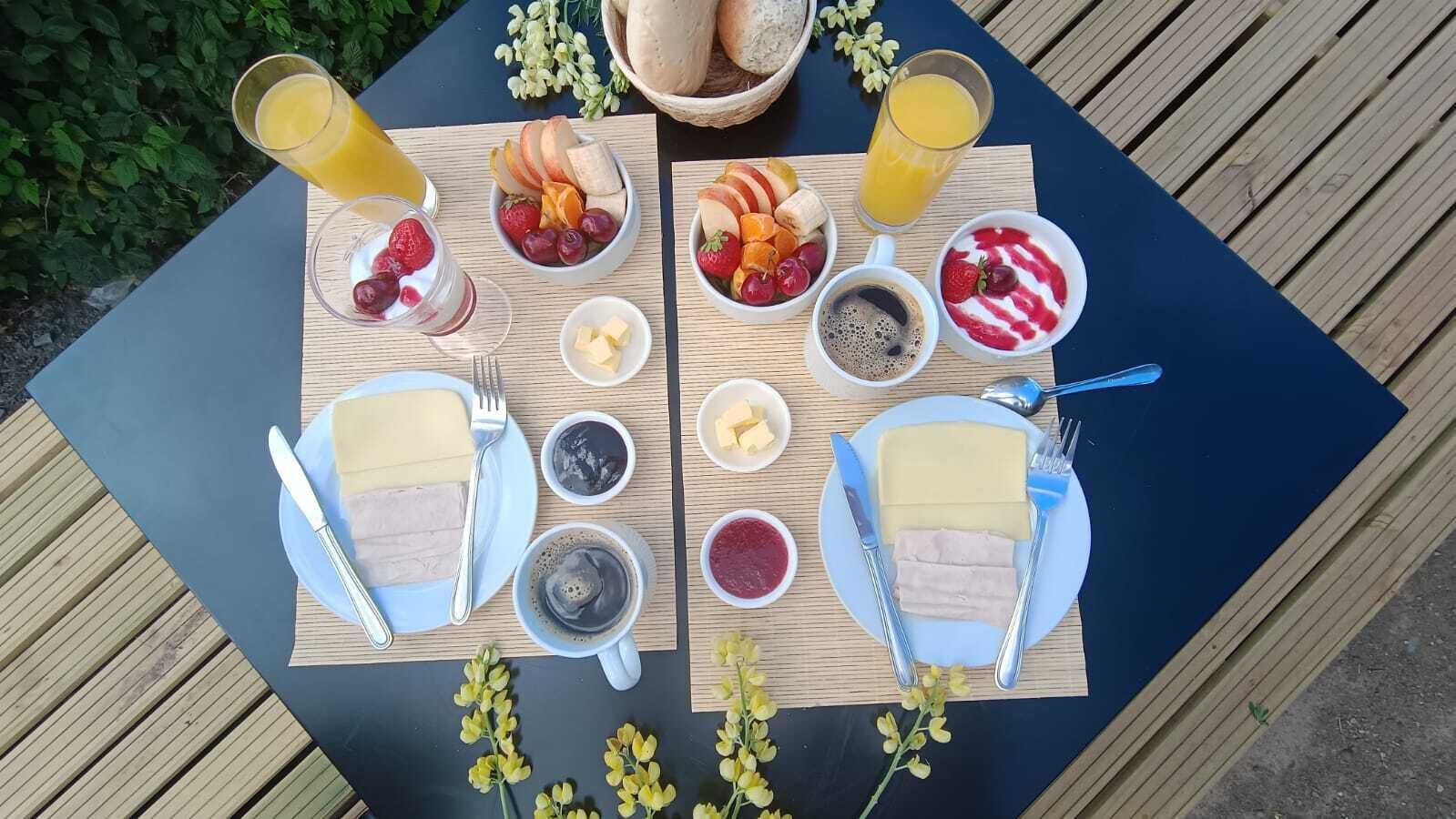 Dos mesas de comedor con alimentos para el desayuno, incluyendo frutas, queso y bebidas, dispuestas al aire libre.