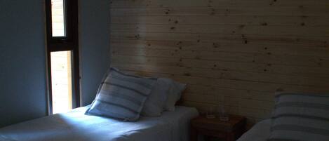 Habitación con dos camas, cada una con ropa de cama a rayas, una mesita de noche de madera y una ventana con persianas.