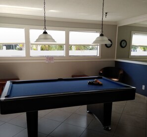 Een pooltafel met twee hanglampen, een klok en een raam met uitzicht op palmbomen.