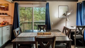 A dining area with a table set for four, a kitchen area with white cabinets, and a view of trees through large windows.