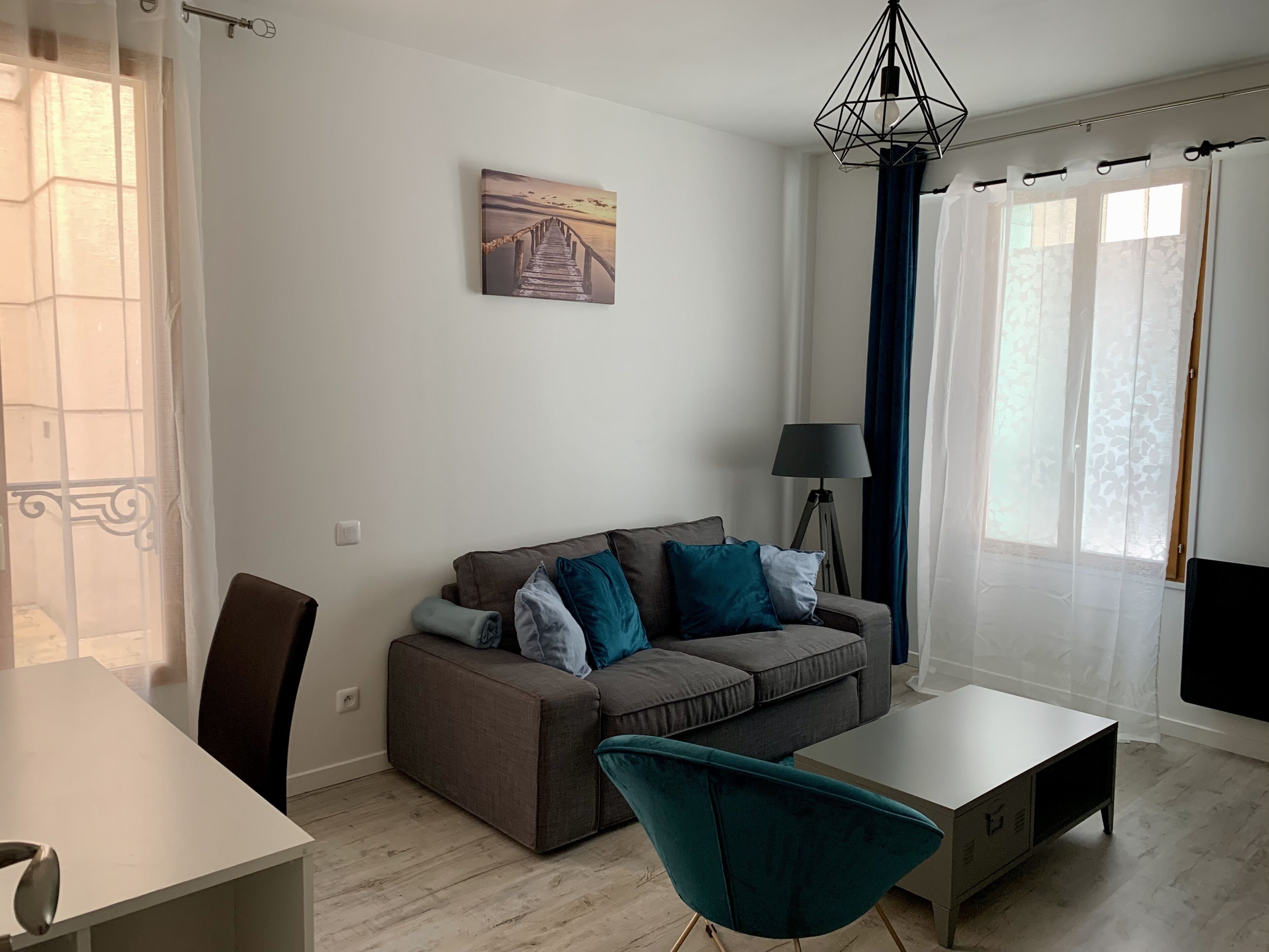 A modern living room with a grey sofa, teal armchair, and a white coffee table. There is a painting of a pier on the wall, a floor lamp, and a window with sheer curtains.