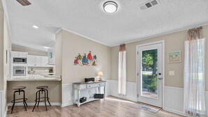 A kitchen with white cabinets, a microwave, and two stools. There is a dining table with a lamp and a wall art piece above it. - Sand Dollars - 5mins to beach, huge lot, pet friendly and boat parking!  (Ocean Isle Beach)