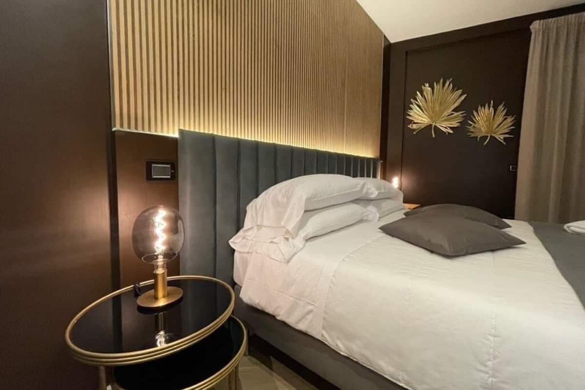 A modern hotel room with a bed, a round table with a lamp, and decorative wall art.