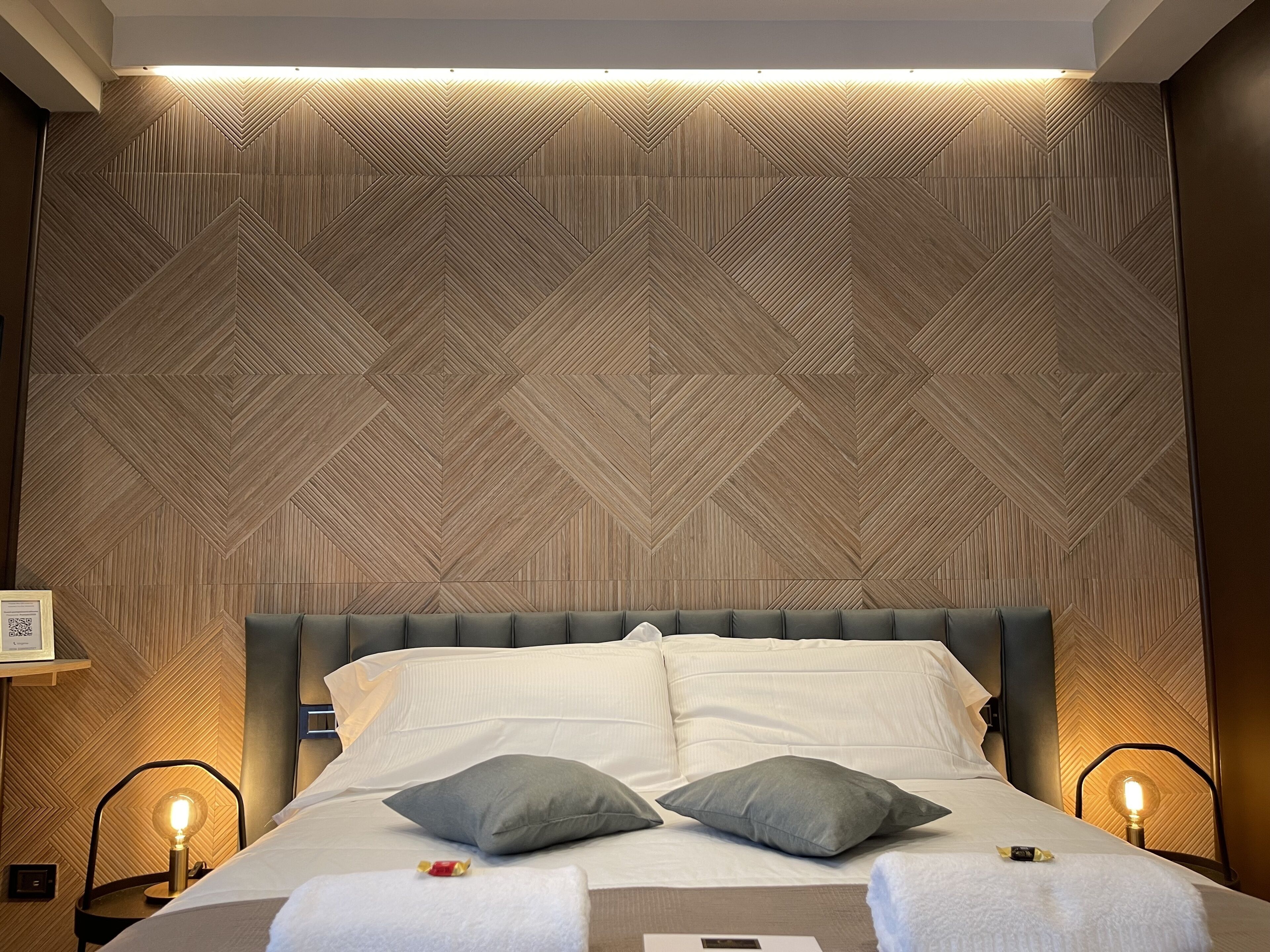 A hotel room with a geometric patterned wall, a bed with white linens, two pillows, and bedside lamps.