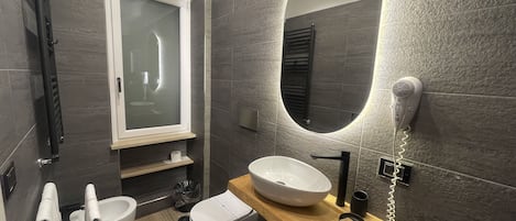 Uma casa de banho moderna com sanita independente, lavatório com bancada de madeira, espelho com iluminação integrada e secador de cabelo fixado na parede.