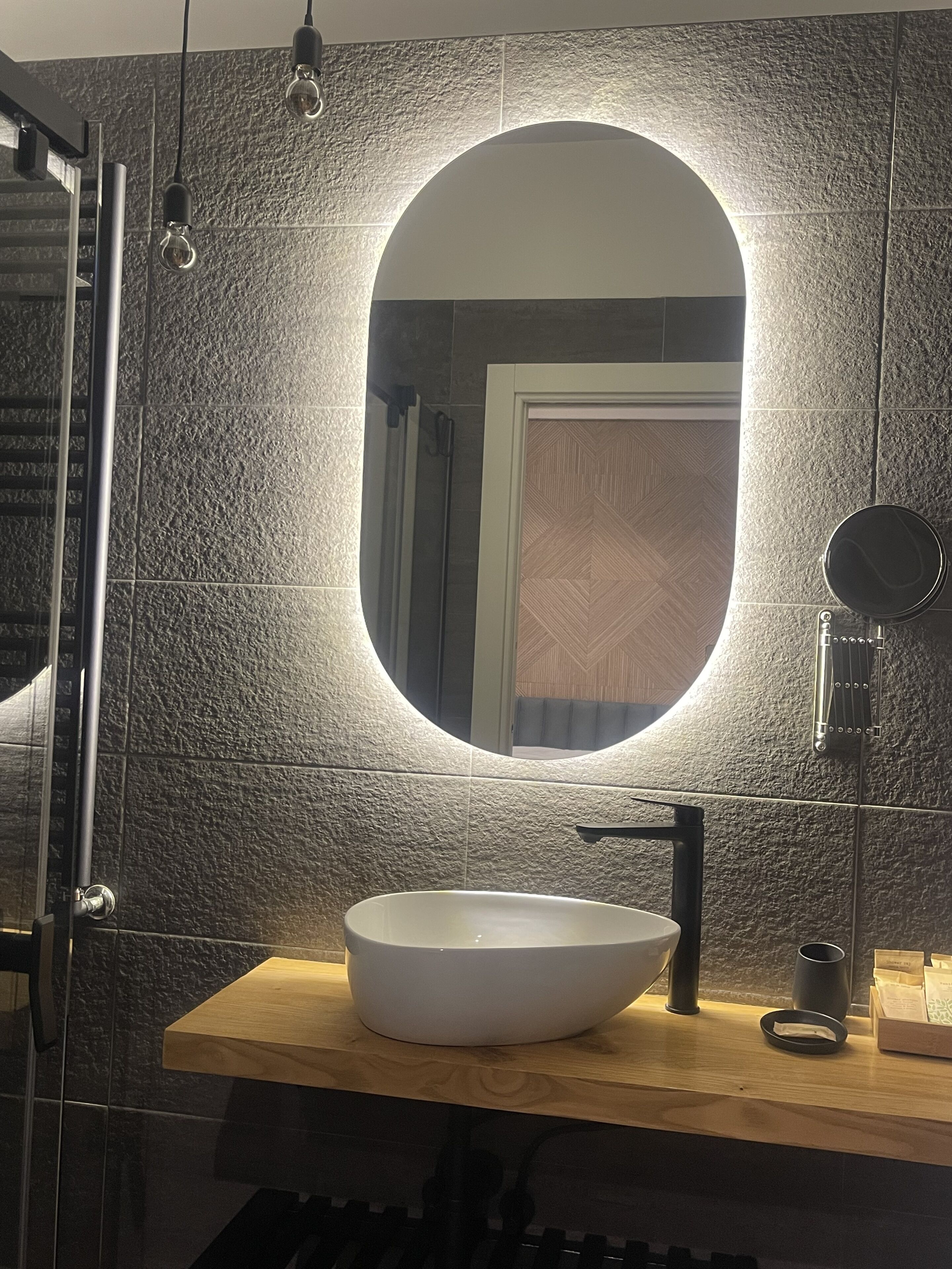 A bathroom with a modern design, featuring a large oval mirror, a vessel sink, and a handheld mirror.