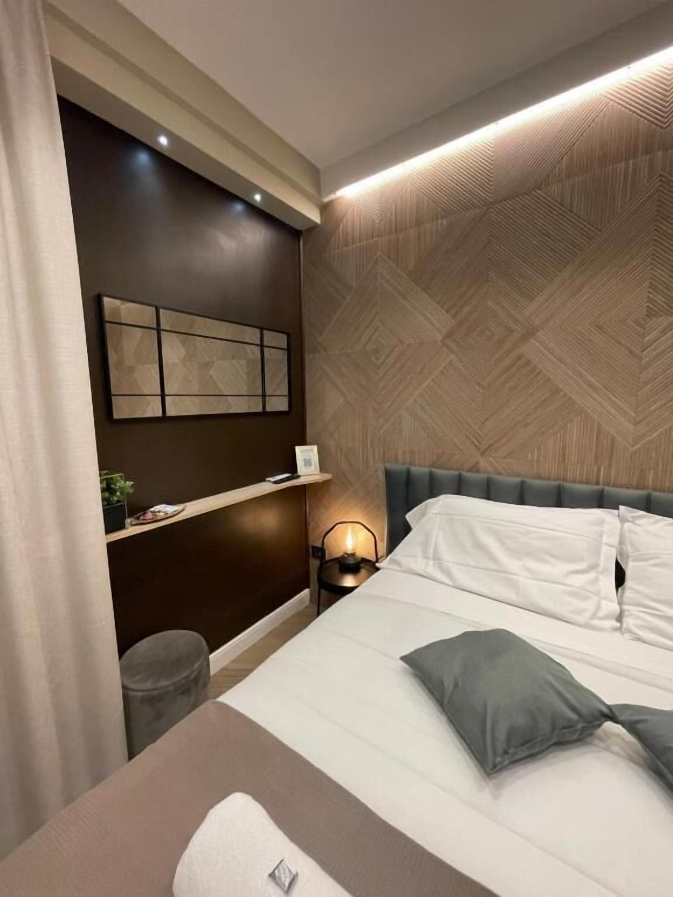 Quarto de hotel moderno com uma cama grande, uma mesa de cabeceira com abajur, uma TV montada na parede e um painel decorativo.