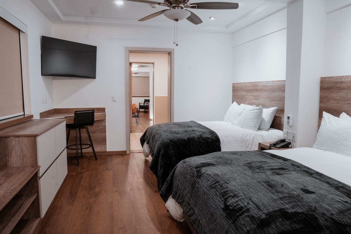A hotel room with two beds, a desk, a chair, a TV, and a ceiling fan.