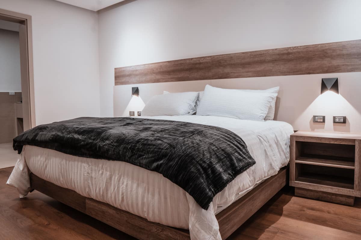 A modern bedroom with a wooden headboard, a large bed with white and black bedding, two bedside lamps, and a wooden nightstand.
