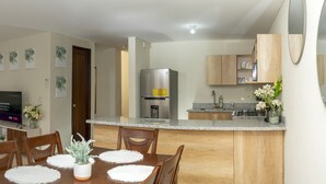 Una sala moderna con un sofá gris, una mesa de comedor con individuales, un televisor y una cocina con nevera y armarios.
