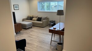 Ein modernes Wohnzimmer mit einer Couch, einem Hocker, einem Couchtisch und einem Schreibtisch mit Lampe. Es gibt ein Fenster mit Jalousien, ein Bild an der Wand und einen Mülleimer.