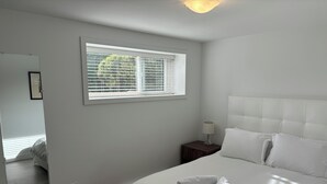 Ein Schlafzimmer mit einem großen Bett, einem Nachttisch mit Lampe, einem Fenster mit Jalousien und einem Spiegel an der Wand.