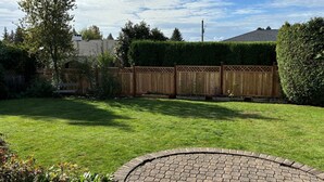 Ein gepflegter Garten mit Holzzaun, gepflasterter Terrasse und verschiedenen Pflanzen und Bäumen.