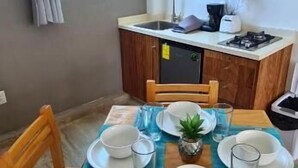 A dining area with a table set for four, a kitchen with a gas stove, and a window with curtains. - Hotel Casa Shula Acapulco (Coyuca de Benitez)