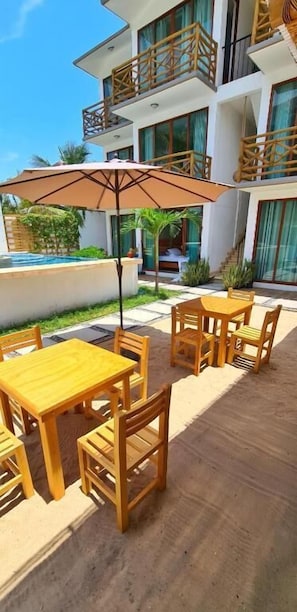A poolside area with wooden tables and chairs, a large umbrella, and a building with balconies in the background.