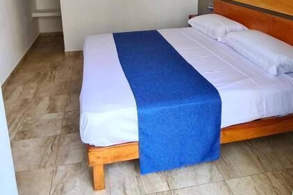 Un dormitorio con cama, manta azul, almohada blanca, cabecero de madera, luz de techo y pared blanca.