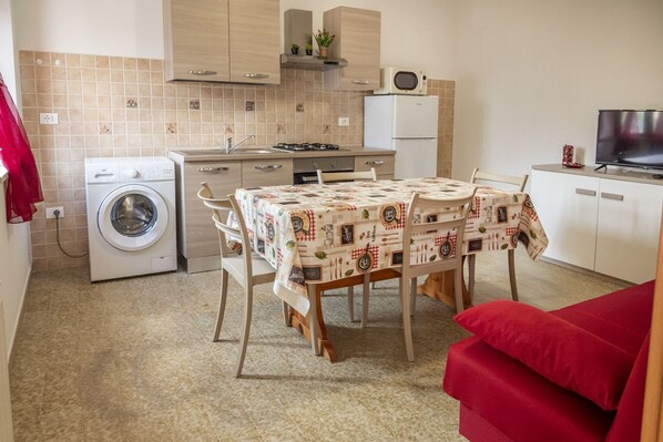 A compact kitchen with a washing machine, a dining table with a patterned tablecloth, and a red sofa.