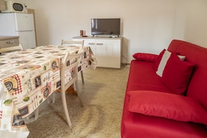 Una camera compatta con un tavolo da pranzo, un divano rosso, un televisore su un mobile basso e un forno a microonde sopra un frigorifero bianco.