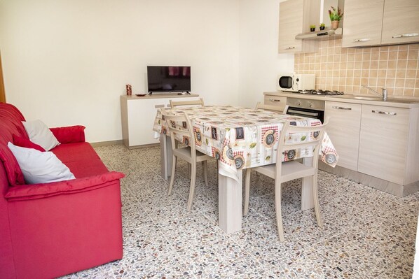 Un espacio habitable compacto con un sofá rojo, una mesa de comedor con mantel estampado, un televisor sobre un mueble, un microondas y un salpicadero de cocina con azulejos.