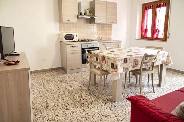 Cocina compacta con microondas, estufa y horno, una mesa de comedor con sillas, y un sofá rojo en una habitación con piso de terrazzo.