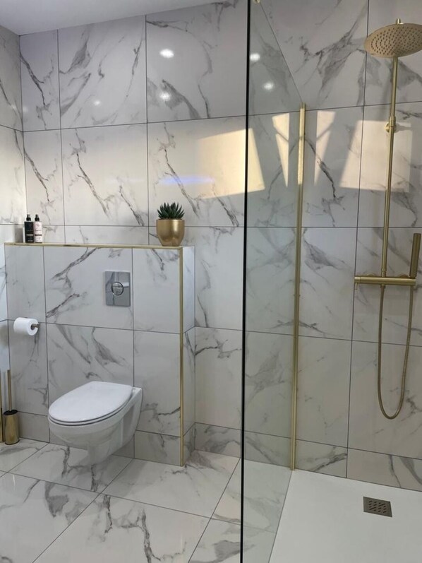 A modern bathroom with marble tiles, a toilet, a shower with a gold showerhead, and a small potted plant on a gold shelf.