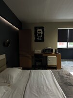 Habitación de hotel con cama, escritorio, refrigerador pequeño y microondas, además de una ventana con persianas.