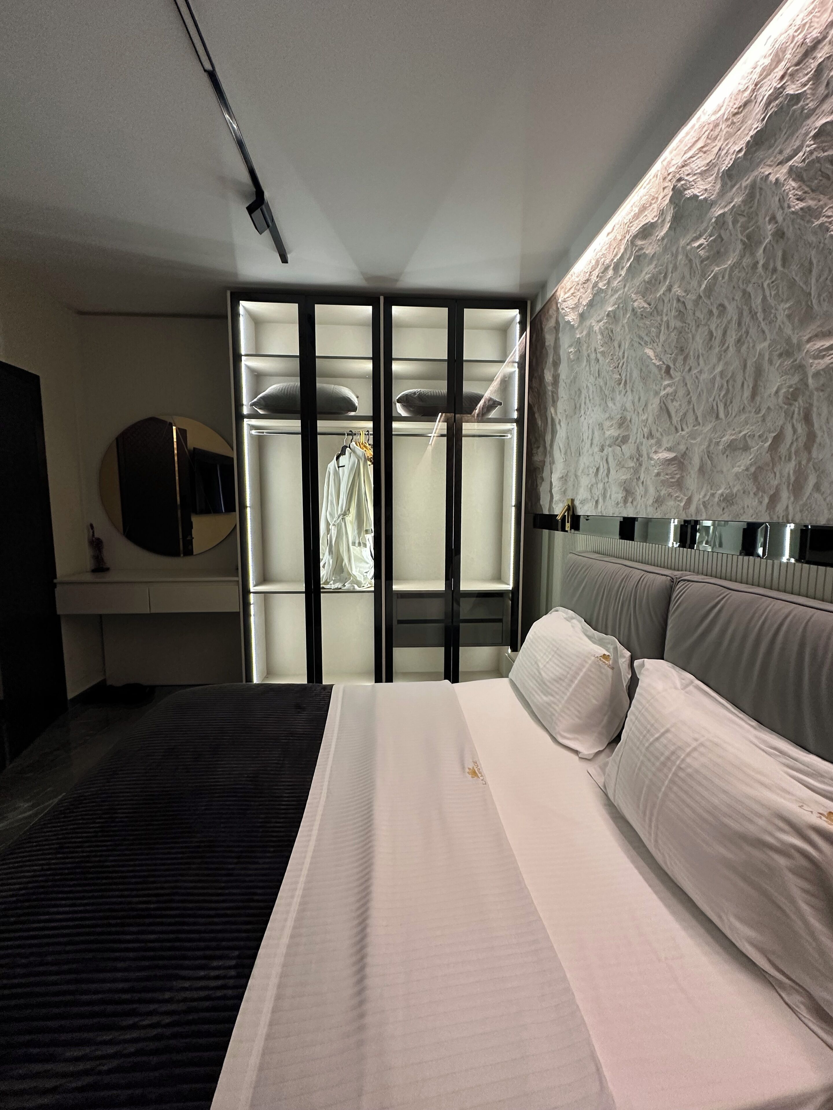 Une chambre d’hôtel moderne dotée d’un grand lit, d’un dressing avec portes coulissantes, d’un grand miroir rond et d’un mur à texture.