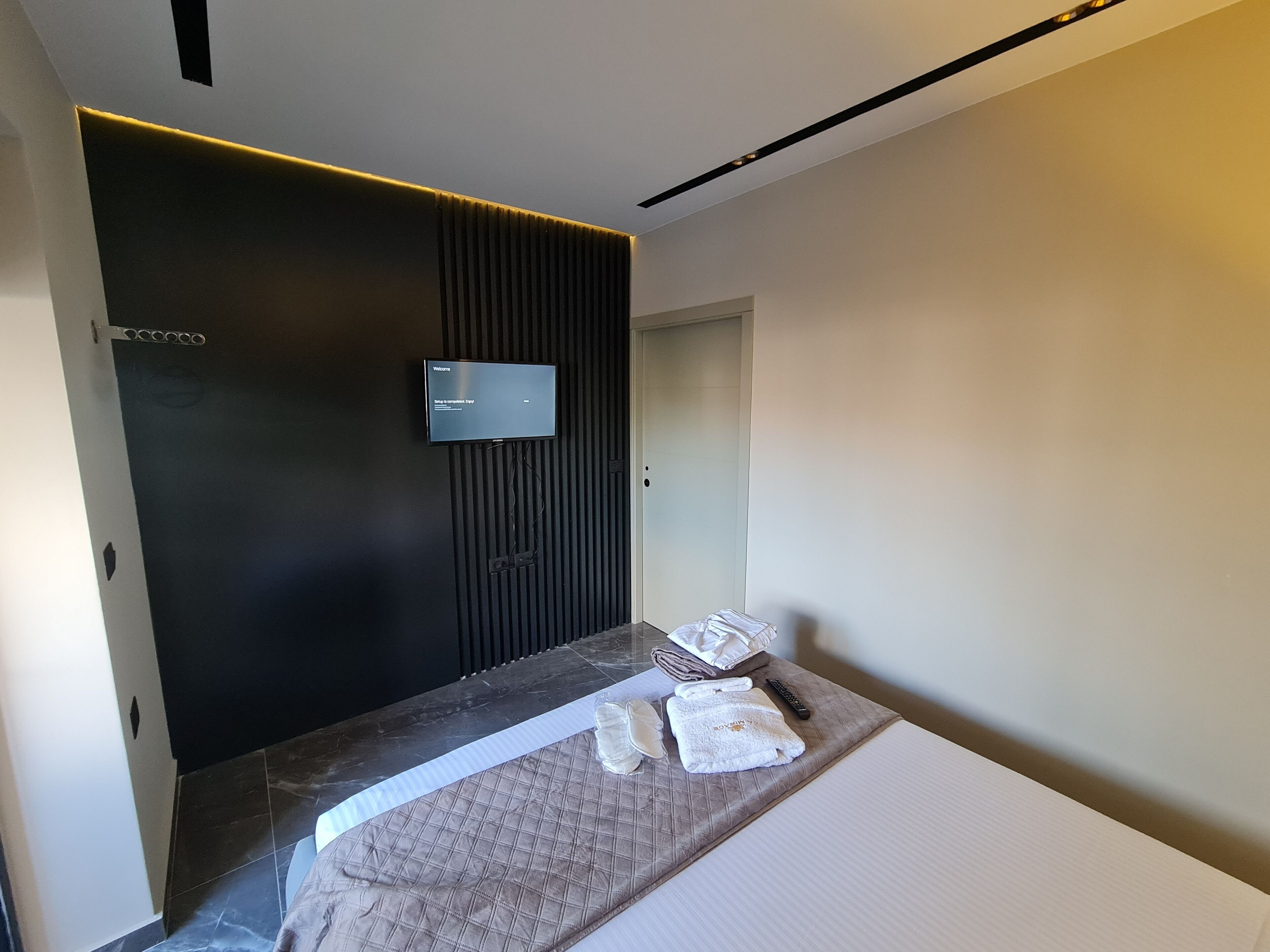 A modern hotel room with a dark accent wall, a flat-screen TV mounted above a bed, and neatly folded towels on the bed.