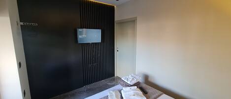 A modern hotel room with a dark accent wall, a flat-screen TV mounted above a bed, and neatly folded towels on the bed.