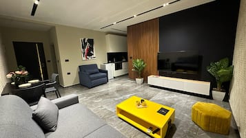 Een moderne woonkamer met een bank, een gele salontafel, een televisie, en een keuken in de achtergrond.