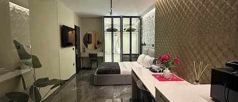 A modern hotel room with a glass partition, a bed, a desk with a computer, and a kitchen area.