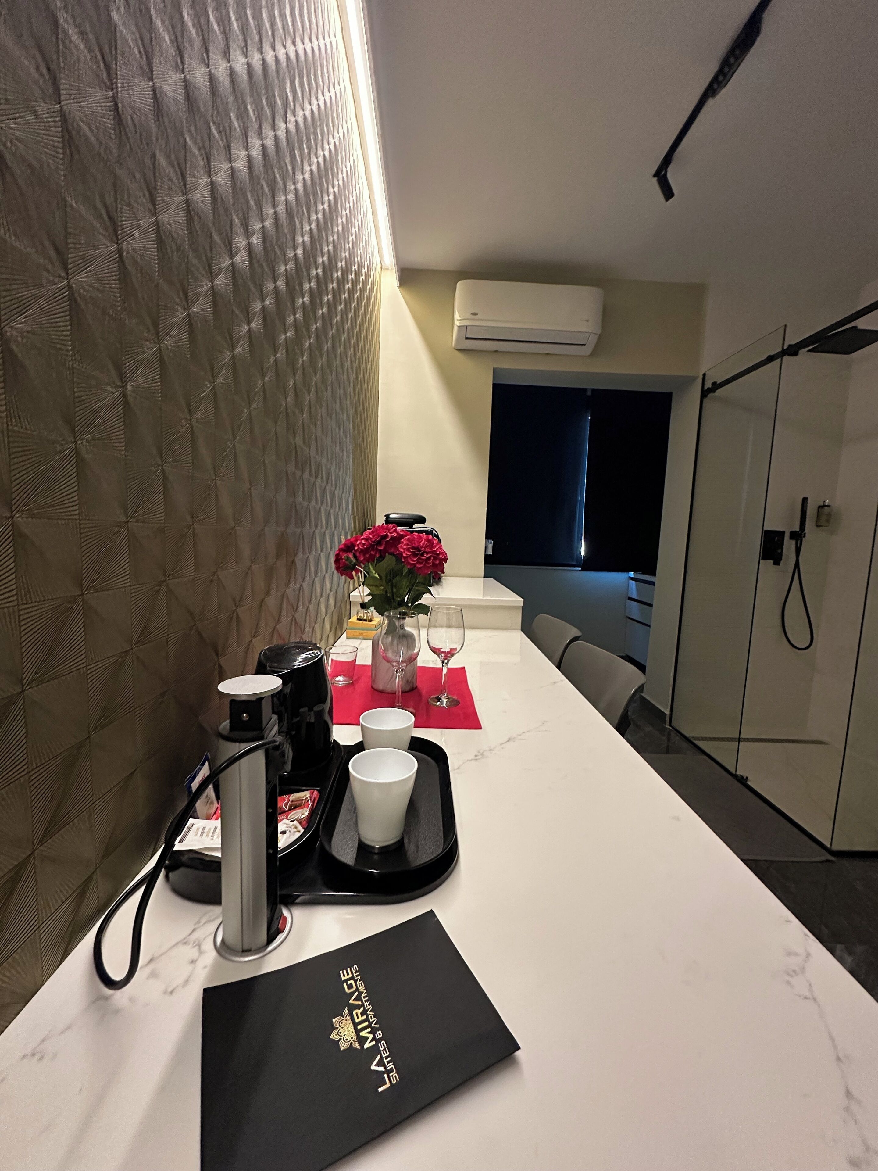A hotel room with a marble countertop, a black tray with a coffee maker, a vase with red flowers, and a black folder labeled "LA MIRAGE".